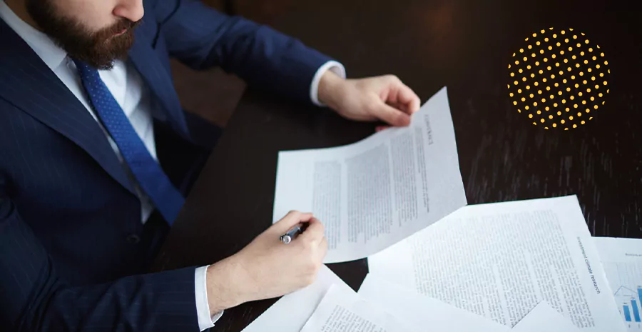 imagem de um homem sentado vestido de terno, segurando um documento e analisando com uma caneta em cima de uma mesa com vários documentos em papeis