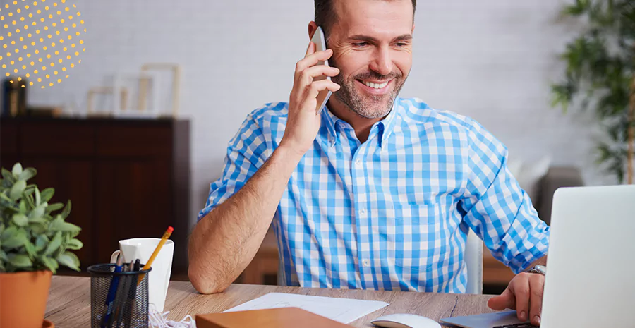 homem com o celular no ouvido olhando para notebook que está em cima de uma mesa de escritório