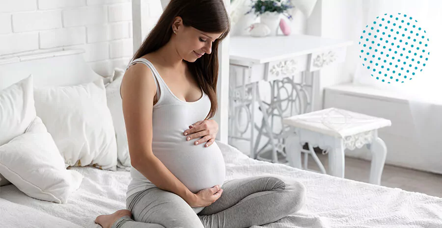 imagem de uma mulher grávida sentada numa cama com as mãos na barriga olhando para a barriga