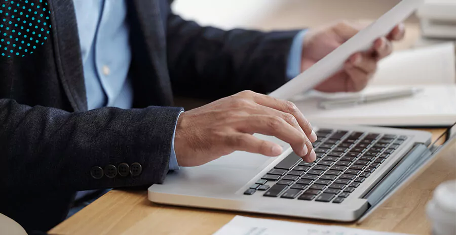 imagem de um notebook aberto sob a mesa, uma pessoa digitando e segurando um papel com a outra mão