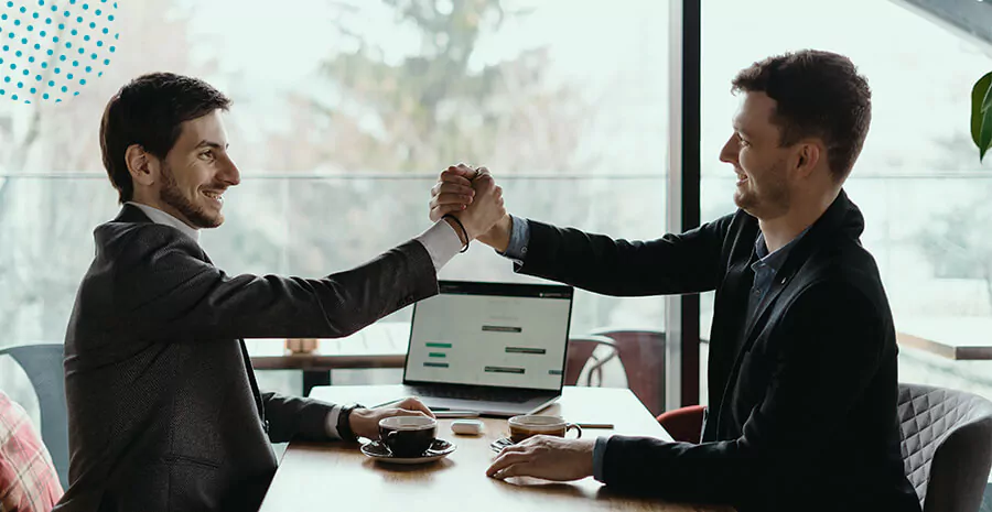 imagem de dois homens sentados, uma mesa entre eles, notebook xícaras de café e os dois apertando as mãos e sorrindo