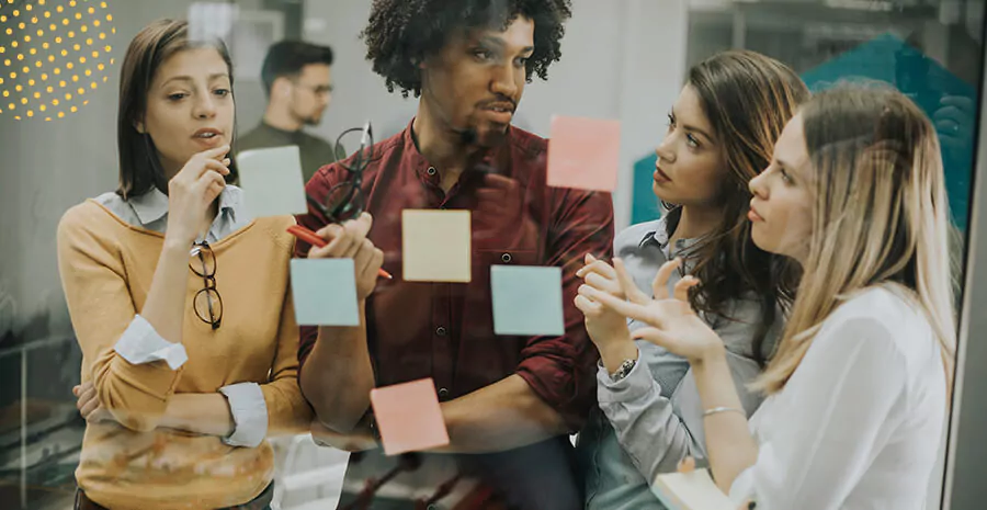 Imagem de um grupo de pessoas, sendo o único homem no centro, duas mulheres ao lado direito e uma ao esquerdo. Eles estão afrente de uma parede de vidro com postits