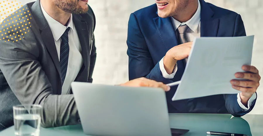 imagem de dois homens de terno sentados conversando em frente uma mesa com notebook e copo de água e um deles mostrando um papel de teste de integridade