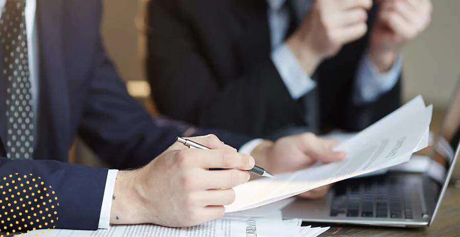 imagem de uma pessoa vestida de terno com um papel e caneta na mão de frente para um notebook numa mesa de trabalho