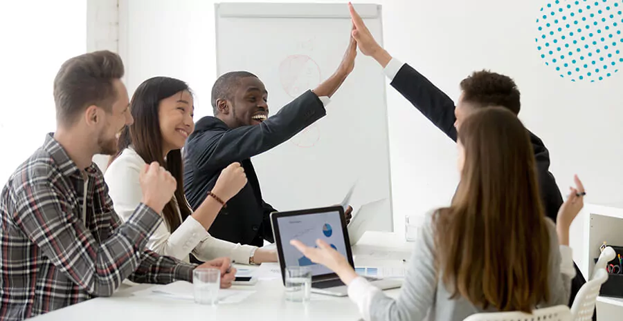 imagem de 5 pessoas sentadas em volta de uma mesa de trabalho e duas tocando as mãos em forma de comemoração