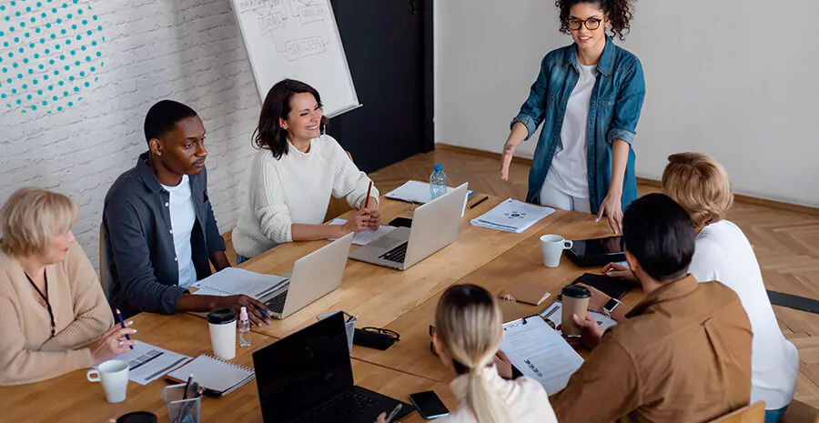 imagem de uma equipe de trabalho em volta de uma mesa e a líder em pé falando com eles