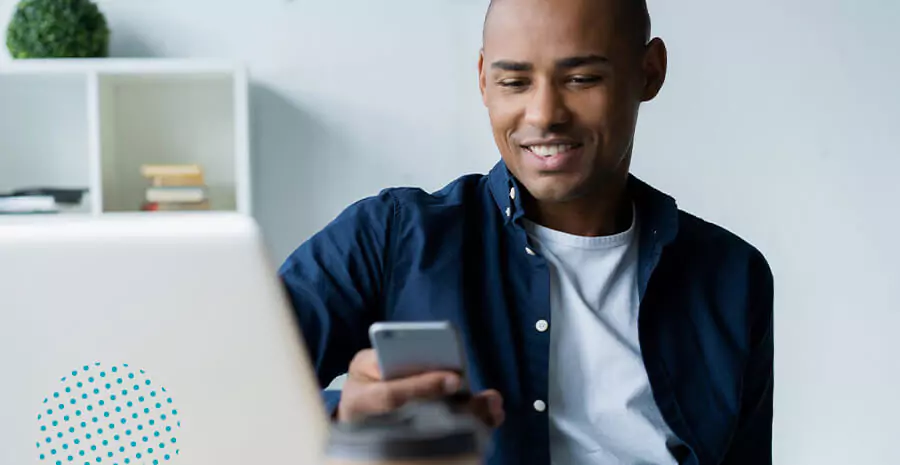 imagem de um homem sentado com um computador na frente, mexendo no celular e sorrindo