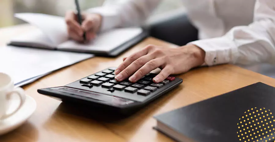 imagem de uma pessoa com a mão em cima de uma calculadora enquanto escreve em um caderno 