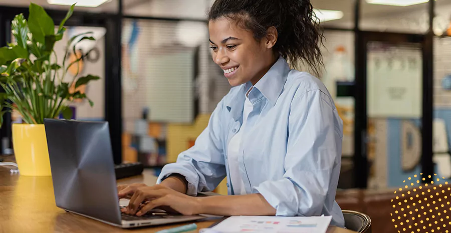 imagem de uma mulher sentada mexendo num notebook em cima de uma mesa com uma planta ao lado trabalhando super feliz