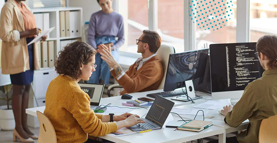 imagem de um equipe de tecnologia em volta de uma mesa com vários computadores e notebook, com três pessoas sentadas mexendo nos computadores e duas em pé