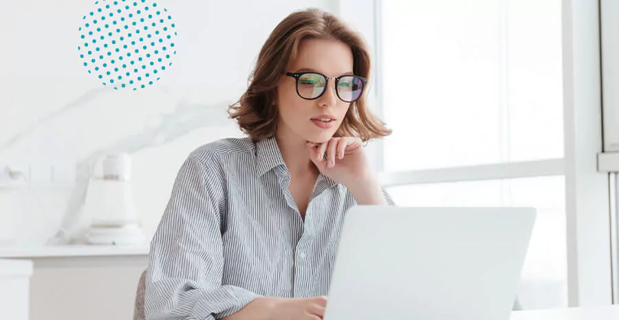 imagem de uma mulher de cabelo curto com óculos sentada com a mão no queixo mexendo num notebook branco a sua frente sob uma mesa