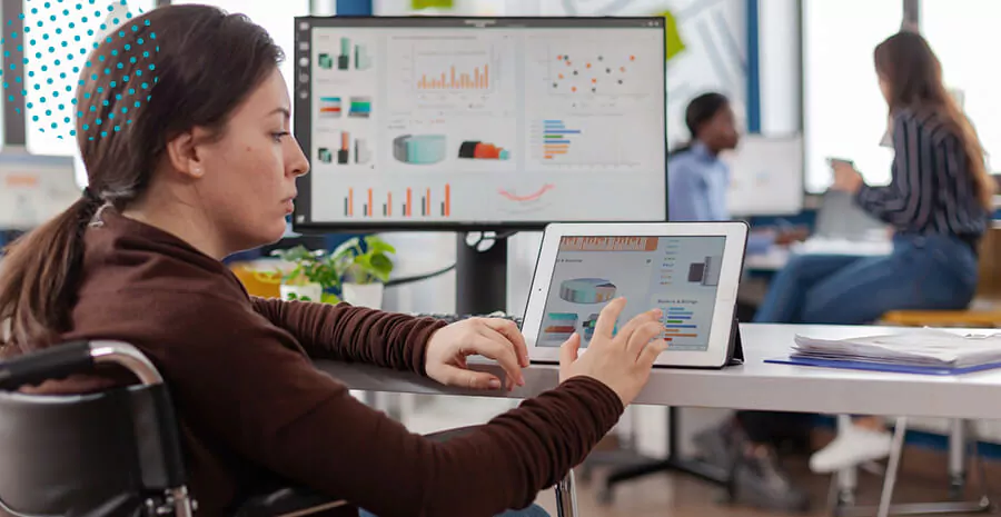 imagem de uma mulher sentada em frente a uma mesa com computador e tablet, mexendo no tablet com gráficos