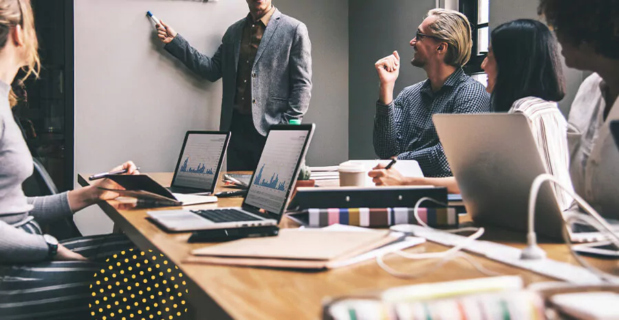 foto de uma mesa com vários notebooks abertos mostrando gráficos de roi e pessoas sentadas em volta com uma pessoa em pé explicando num quadro branco