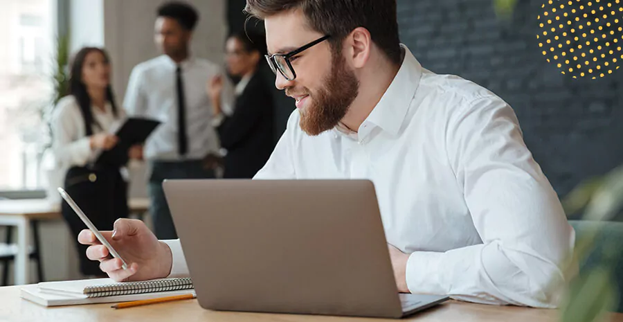 imagem de um homem segurando um celular olhando para ele e com um computador na frente