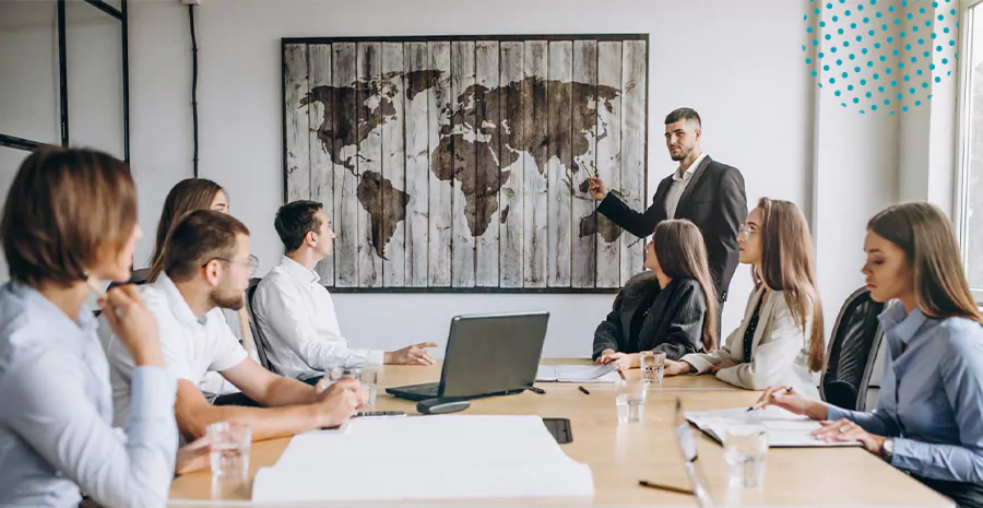 imagem de um grupo de pessoas sentado ao redor de uma mesa olhando um homem em pé apontando para um mapa mundi