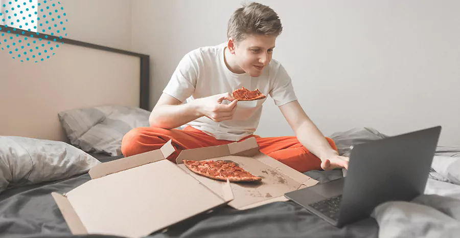 imagem de um homem sentado na cama comendo pizza e mexendo no computador