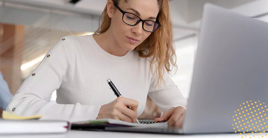imagem de uma mulher concentrada sentada na frente de um computador e escrevendo em um caderno