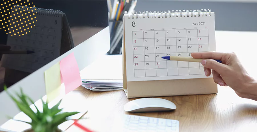 imagem de uma pessoa apontando uma caneta para um calendário em cima de uma mesa ao lado de um mouse, um teclado e folhas 