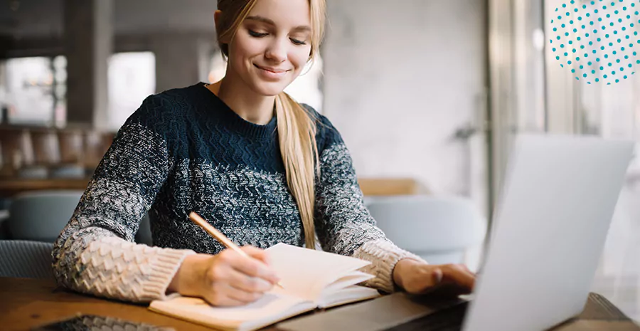 imagem de uma mulher sorrindo sentada na frente de um computador e escrevendo em um caderno