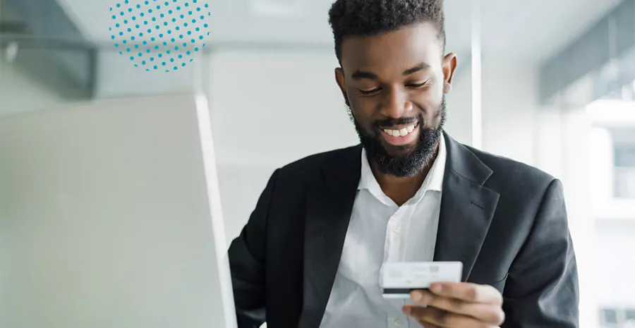 imagem de um homem sorrindo sentado na frente de um computador e segurando um cartão de crédito