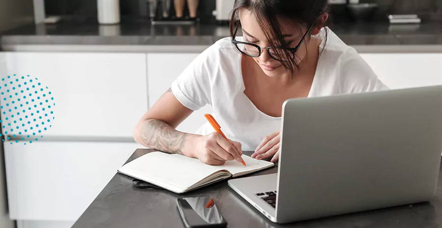 imagem de uma mulher sentada na frente de um computador e escrevendo em um caderno