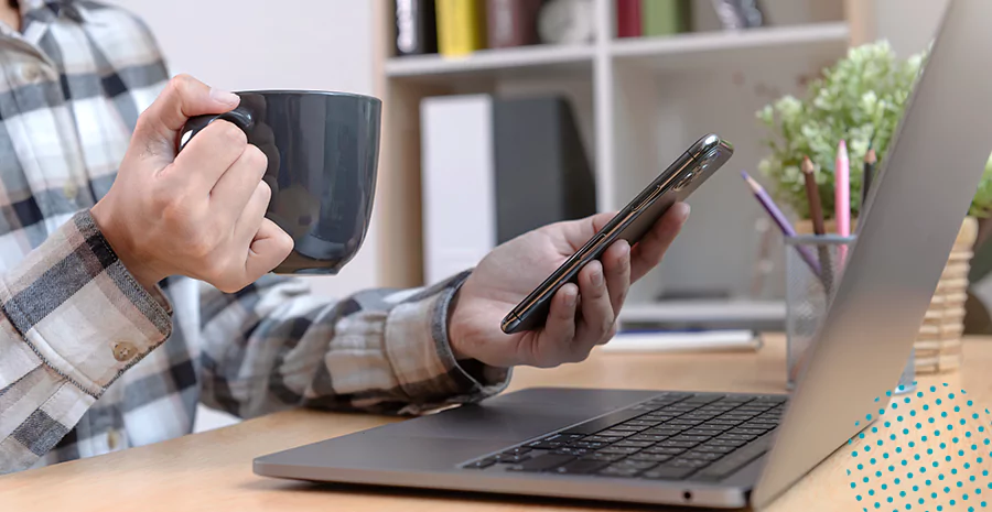 imagem de um homem sentado na frente de um computador segurando uma xícara e um celular