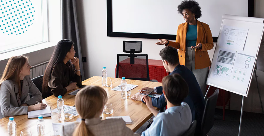 imagem de cinco pessoas sentadas ao redor de uma mesa olhando para uma mulher em pé na frente deles apontando para um quadro branco