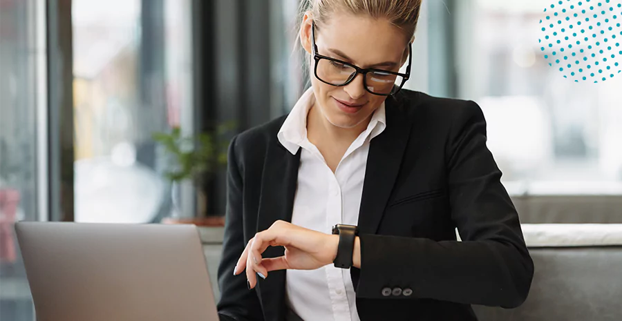 imagem de uma mulher sentada na frente de um computador e olhando para seu relógio de pulso