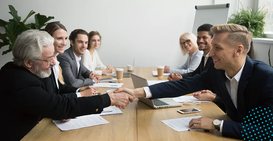 imagem de um grupo de pessoas sentado ao redor de uma mesa em um escritório com dois homens se cumprimentando