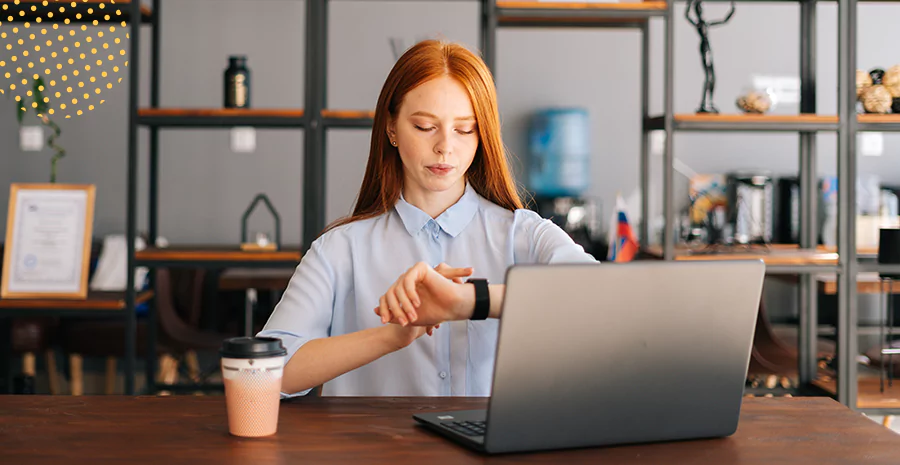 Imagem de uma mulher olhando para o relógio sentada em um escritório com um computador e um copo em cima da mesa