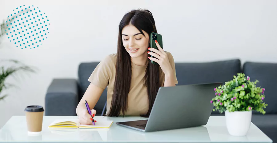 imagem de uma mulher sentada na frente de um computador falando ao celular e escrevendo em um papel