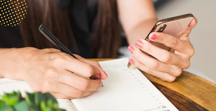 Mulher utilizando um celular enquanto escreve em um caderno