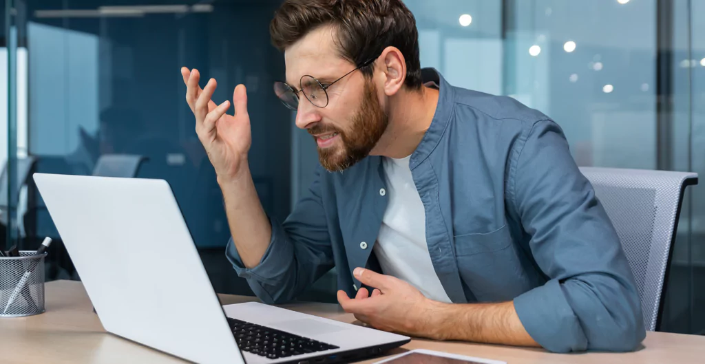 homem em frente ao  notebook com expressão de irritado 