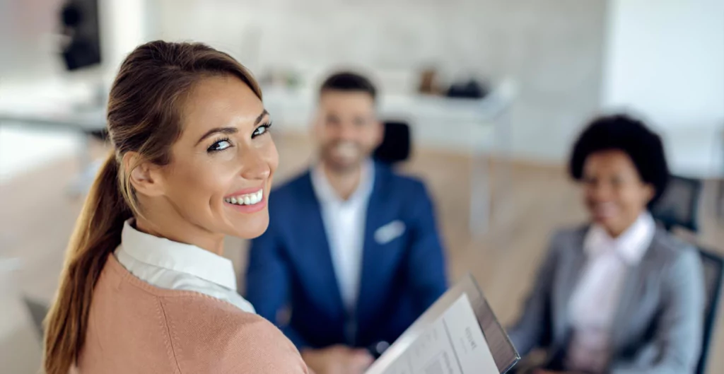 uma mulher sorrindo olhando para a câmera e duas pessoas sentadas atrás desfocadas