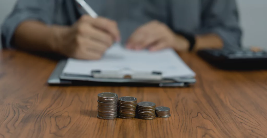 Um homem está escrevendo em um pedaço de papel  ao fundo e na frente  moedas na mesa