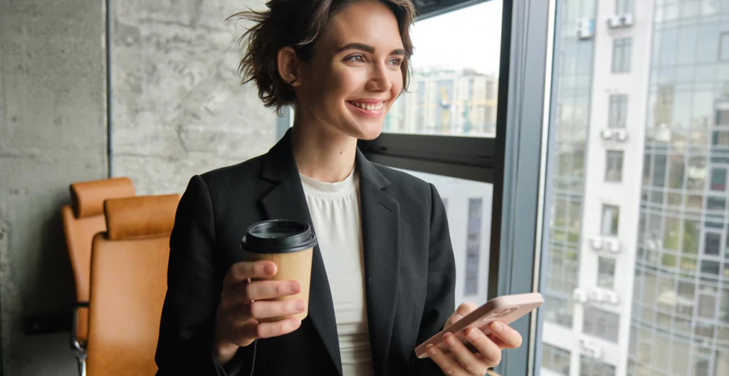 mulher sorridente segurando um copo de café e o celular