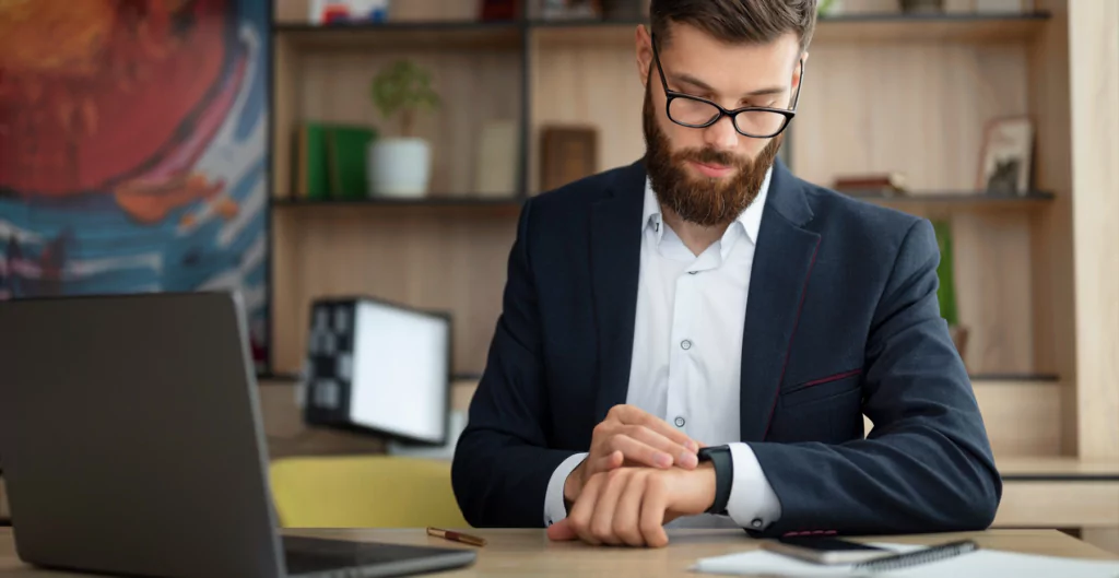 homem sentado de frente a um notebook olhando para o relógio de pulso