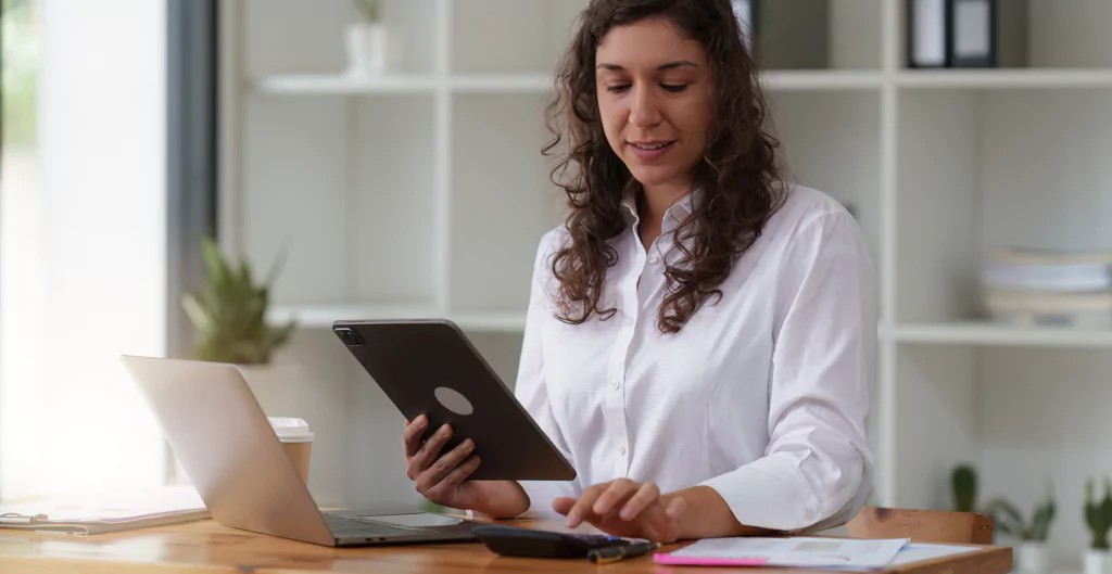muher segurando um tablet e sentada de frente para um notebook e uma calculadora