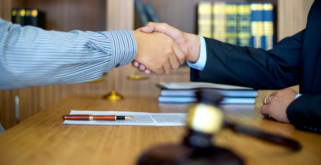 duas pessoas se cumprimentando com um aperto de mão em cima de uma mesa com um contrato e uma caneta