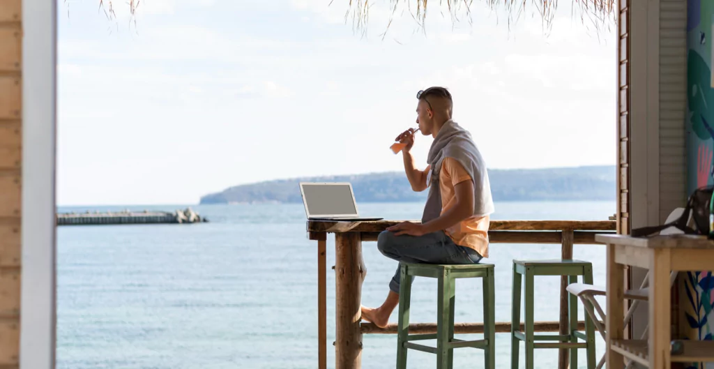 homem sentado de frente para um notebook em uma praia com uma bebida na mão
