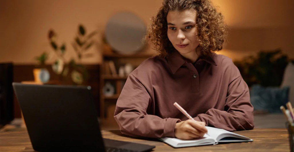 imagem mostra mulher sentada de frente para o notebook e escrevendo em um caderno