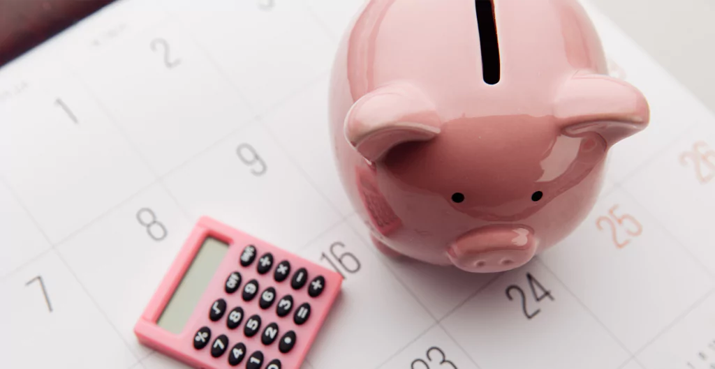 Cofrinho e calculadora sobre calendário, simbolizando planejamento para o décimo quarto salário.