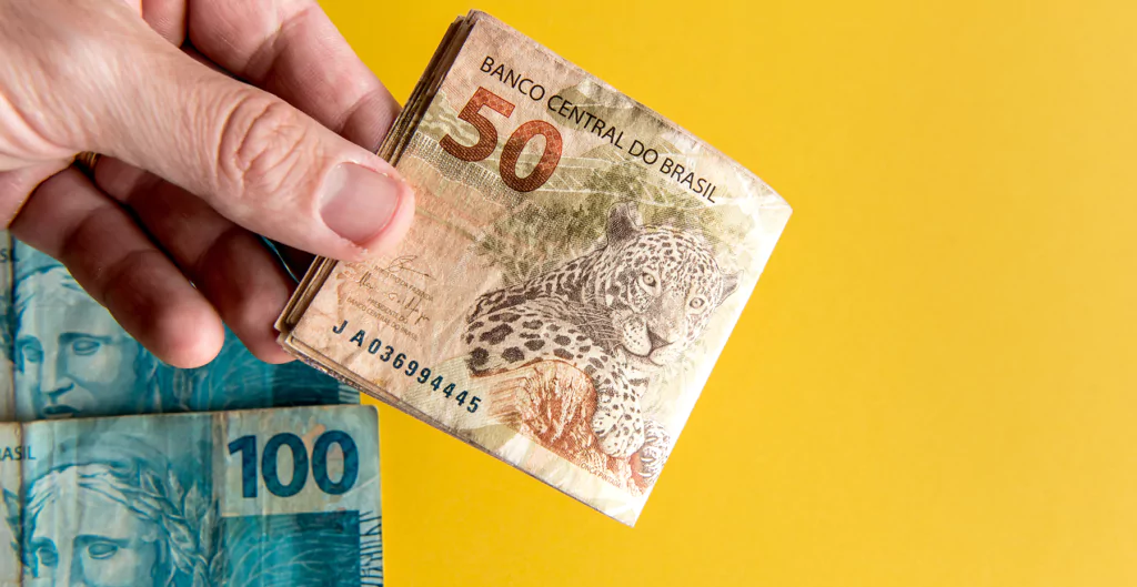 Mão segurando cédulas de 50 e 100 reais, ilustrando pagamento do décimo quarto salário.