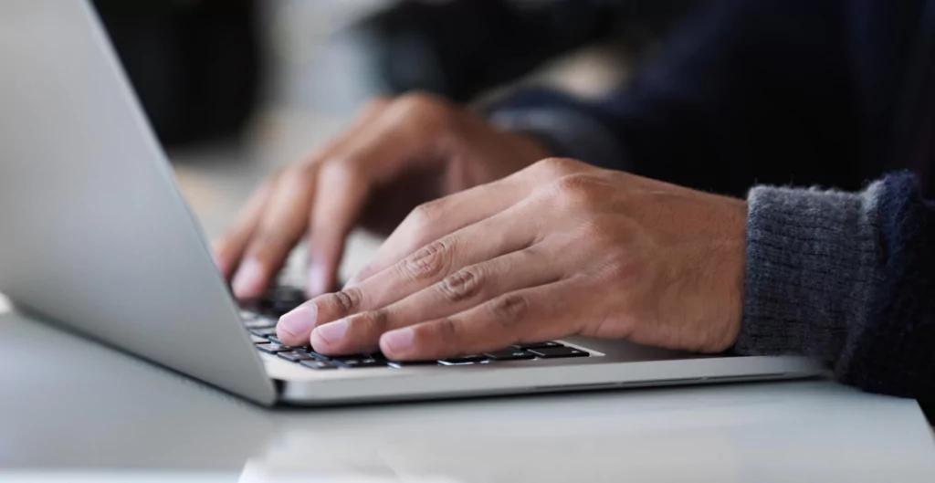 Visão aproximada de mãos masculinas digitando em um teclado de notebook prata.