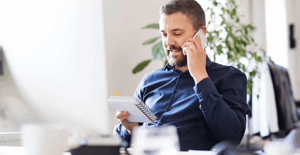 Homem de negócios falando ao telefone, segurando um caderno, em ambiente de escritório moderno com plantas ao fundo.