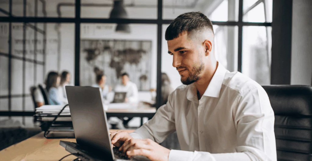 homem sentado digitando em notebook, mas com sala de reunião ao fundo.