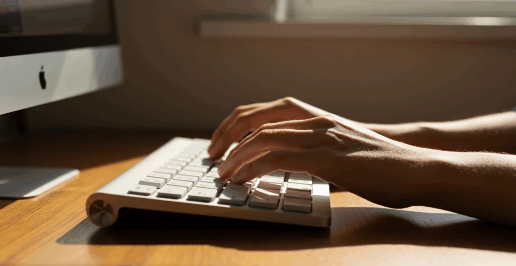 Pessoa digitando em um teclado de computador, ambiente de escritório com iluminação natural.