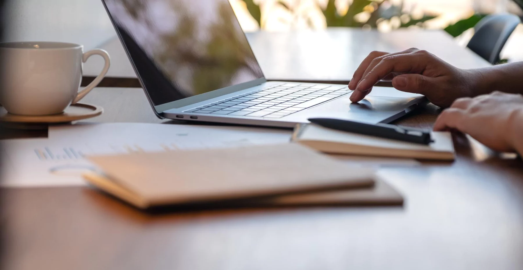 Pessoa trabalhando em um laptop em um ambiente de escritório com livros, uma xícara de café e uma vista de janelas com plantas ao fundo, ideal para quem busca dicas de produtividade com tecnologia.