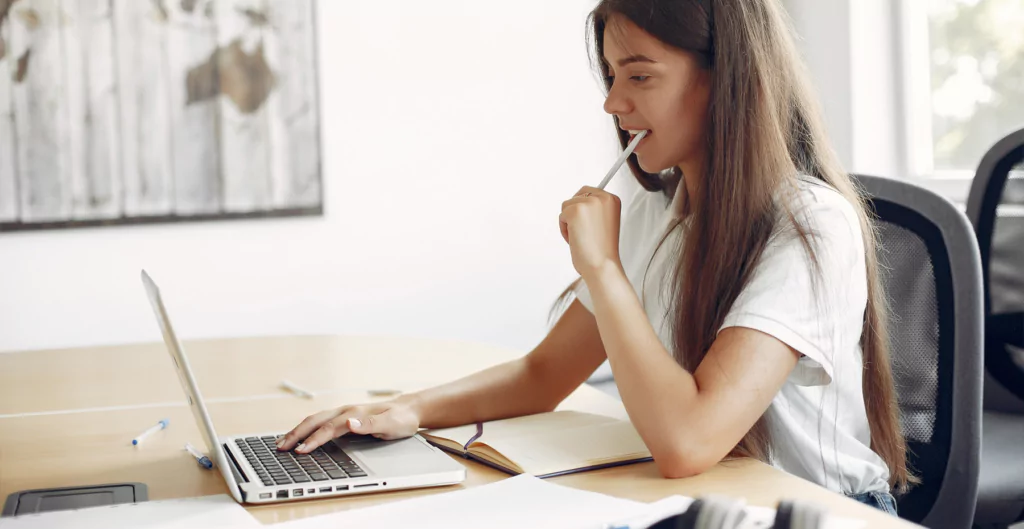 mulher em um escritório sentada em frente a um notebook e com uma caneta na boca