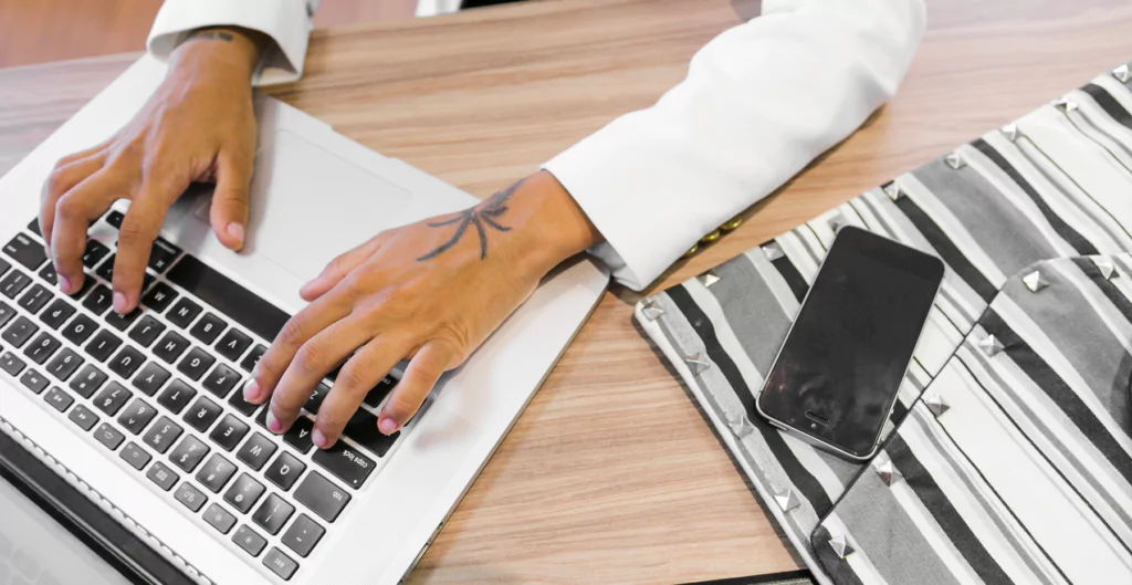 imagem mostra uma mão mexendo no notebook que está em cima da mesa e do lado de um celular